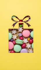 Creative flat lay of a gift box shape cut from yellow paper revealing colorful macarons, cookies and sweets