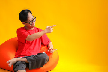 Student Boy with Glasses Pointing Excitedly While Sitting on Beanbag Isolated on Yellow background