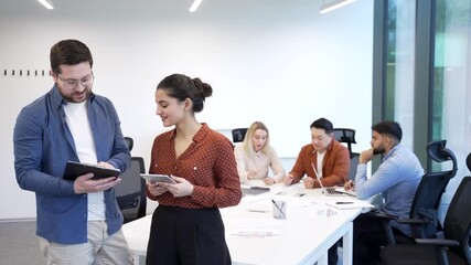 Two co-workers, man and woman engaged in discussion holding a digital tablet standing on background diverse team employees in business office. Teamwork and collaboration. Friendly positive cooperation - Powered by Adobe
