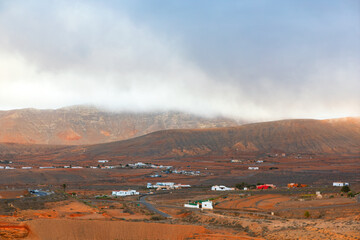 Typical Canary Islands landscape with rugged brown mountains shrouded in low clouds, creating dramatic and misty atmosphere. Scattered white houses dot arid terrain of picturesque rural scenery