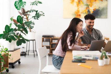 A young woman and man collaborate on laptops at a desk, surrounded by plants and art, in a bright, modern workspace.