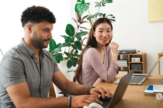 A man and a woman work together on laptops at a wooden desk, surrounded by plants and office supplies, fostering a collaborative and productive atmosphere. - Powered by Adobe