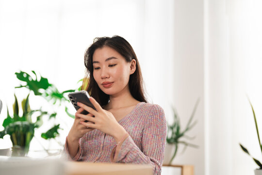A young woman with dark hair is looking at her smartphone while sitting indoors. She is wearing a colorful knitted top.