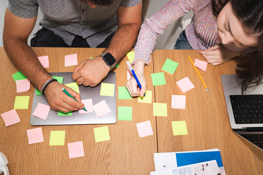 Two people collaborate on a project, using sticky notes and a laptop to brainstorm ideas and sketch concepts on a wooden table.