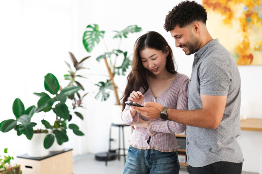 A smiling couple shares a moment looking at a mobile phone together in a bright, plant-filled room.