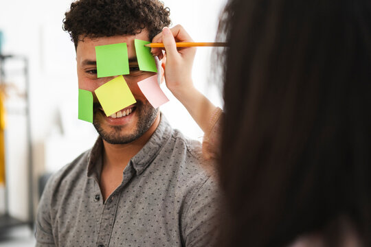 A person writes on sticky notes attached to another person's face, symbolizing brainstorming or a creative session.