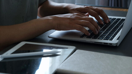 Man typing on laptop with tablet and stylus on desk, working in low light environment, focused and productive atmosphere for remote work or creative project