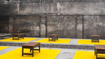 Minimalist outdoor seating area with small wooden tables placed on bright yellow mats against a raw...