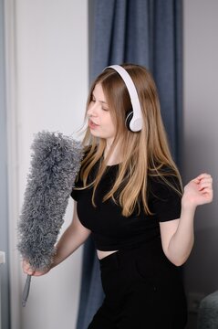 Woman dances and sings while cleaning house, holding pipidaster and listening to music on wireless headphones. Concepts of order and cleaning.