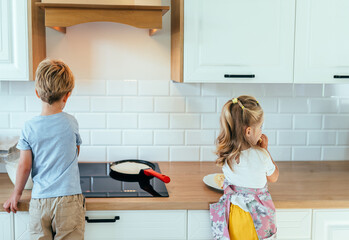 Two children seen from behind cooking pancakes on stove in kitchen