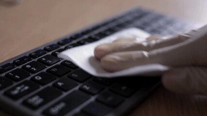 A hand in a glove cleans a computer keyboard with a disinfectant wipe. Close up of sanitizing office equipment for hygiene and disease prevention. - Powered by Adobe