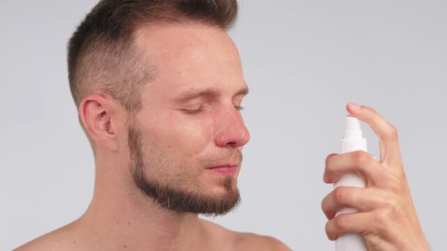 Man uses a cosmetic spray, hydrating facial mist as part of skincare routine. Young adult man applying the cooling mist, spraying facial toner to his face on neutral background. Skincare for man.