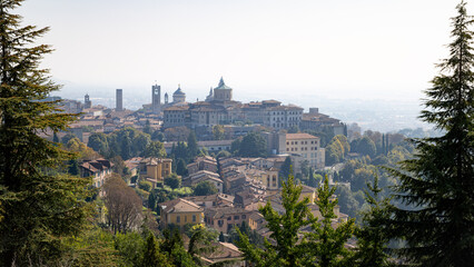 A panoramic view of Bergamo with numerous buildings and a hill in the background. The sky is overcast and the sun is shining.