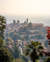 A panoramic view of Bergamo with numerous buildings and a hill in the background. The sky is overcast and the sun is shining.