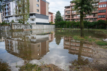 Floodwater blankets an urban area, mirroring buildings, trees, and sky in a striking reflection...