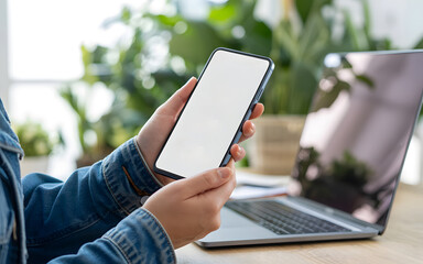 Person holding smartphone with blank white screen near laptop, representing mobile technology, app design, website mockup, and digital communication concept