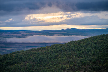Mist drifts through a sunlit valley beneath forested hills and a dramatic, cloud-streaked sky.