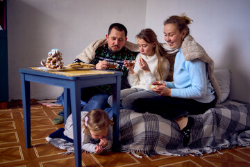 a family with daughters toddler and teenager cozy together drinking tea and eating gingerbread Christmas tree, naughty toddler hiding under the table