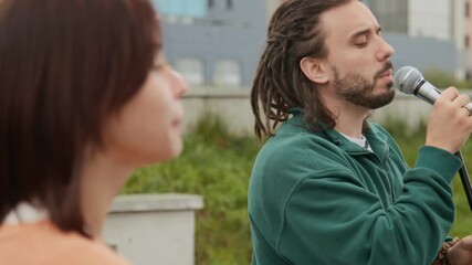 Medium close-up of young Caucasian male Caucasian rock band singer performing new hit with female bandmate accompanying on electric guitar while busking in city street in summer