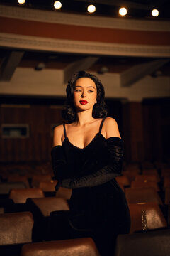 Woman in black dress stands alone in an empty theater with vintage wooden seats during a quiet night