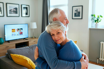 Senior Caucasian woman embracing senior Caucasian man in living room, woman smiling at camera while man looking away, couple standing close together in affectionate pose