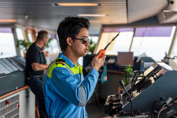 A male officer in a bright yellow and blue safety uniform is on the navigational bridge of a merchant ship, speaking into a handheld radio.