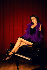 Performing artist in a purple feather boa poses on a grand piano with red curtains in the background during a glamorous evening event