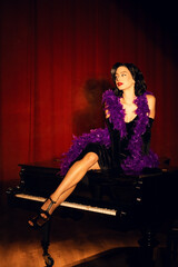 Performing artist in a purple feather boa poses on a grand piano with red curtains in the background during a glamorous evening event