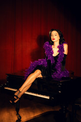 Performing artist in a purple feather boa poses on a grand piano with red curtains in the background during a glamorous evening event