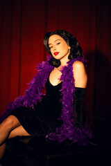 Performing artist in a purple feather boa poses on a grand piano with red curtains in the background during a glamorous evening event