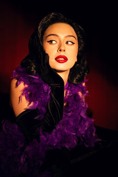Performing artist in a purple feather boa poses on a grand piano with red curtains in the background during a glamorous evening event
