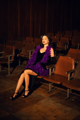 Elegant woman in black dress with purple feather boa sits in empty theater during evening event
