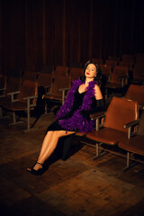 Elegant woman in black dress with purple feather boa sits in empty theater during evening event