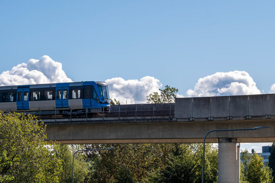 Modern elevated metro train crossing a concrete bridge surrounded by trees and blue sky with bright clouds over Stockholm, showing integration of transport and urban architecture