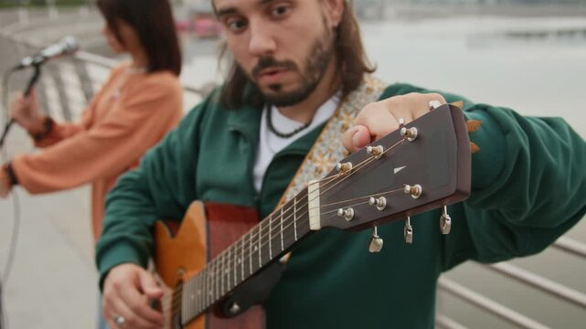 Medium close-up of young Caucasian man turning pegs on acoustic guitar, tuning strings, while female companion is setting up microphone on stand for improvised street concert