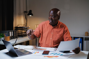 African American businessman working remotely from a home office, having a late-night video call, discussing and analyzing financial documents and charts with a pen