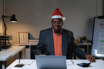 Mature African American businessman wearing a Santa hat while sitting at his office desk, participating in a video call on his laptop during the holiday season