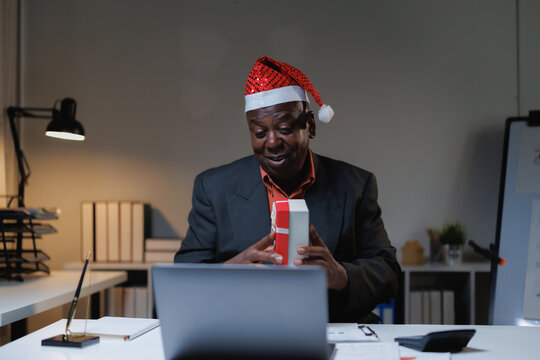 Black businessman wearing a santa hat and smiling while opening a christmas gift during a virtual video call holiday celebration from his office, connecting with colleagues online