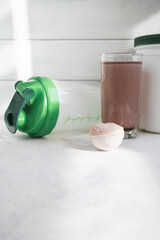 Scoop of whey protein powder, chocolate protein shake in glass, shaker bottle and white plastic protein jar on light surface, fitness nutrition concept
