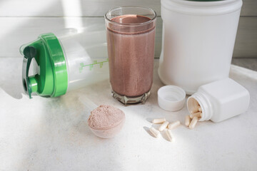 Scoop of whey protein powder, chocolate protein shake in glass, white capsules, shaker bottle and white plastic protein jar on light surface, fitness nutrition concept