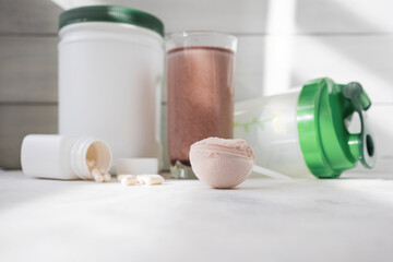 Scoop of whey protein powder, chocolate protein shake in glass, white capsules, shaker bottle and white plastic protein jar on light surface, fitness nutrition concept