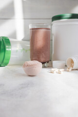 Scoop of whey protein powder, chocolate protein shake in glass, white capsules, shaker bottle and white plastic protein jar on light surface, fitness nutrition concept