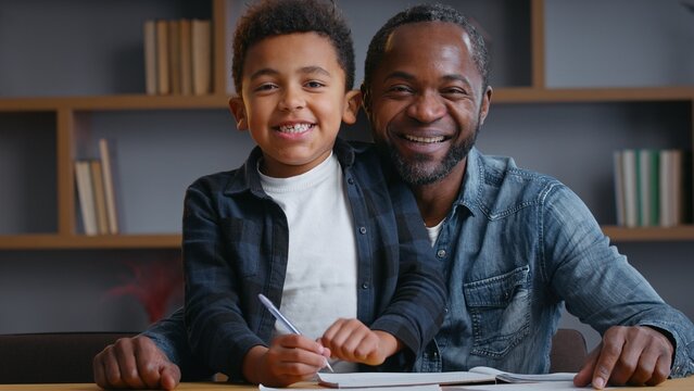 African American family smiling portrait study looking at camera ethnic father dad with little son kid child boy studying learning at table writing homework distant education video call positive look