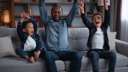 African American happy family watching TV rejoice winning sport game yelling celebrating father with two sons boys kids children watch football championship television match at home having fun laugh