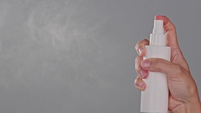 Close-up of a hand spraying a mist from a white bottle on a gray background. Hair spray or heat protection in use with copy space. Haircare product advertising concept.