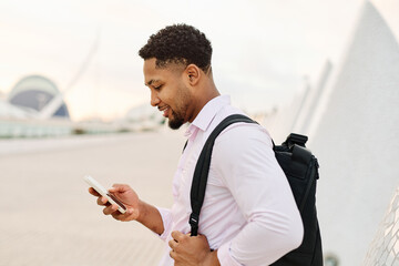 Portrait of a young businessman man using a smartphone mobile phone walking down the street, surrounded by moder corporate office buildings and modern city architecture