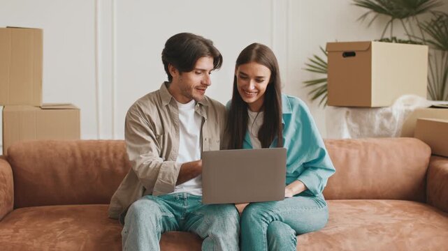 A young couple sits together on a cozy sofa, reviewing real estate listings on a laptop amidst moving boxes in their new apartment. They appear happy and excited about their relocation.