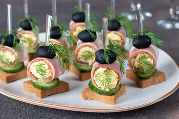 Canapes with bacon, cucumber and black olives on croutons on white plate