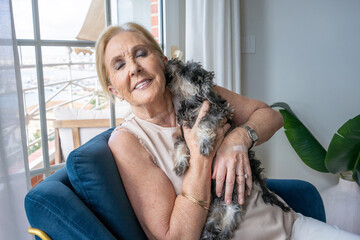 A smiling senior woman hugging with tenderness her schnauzer puppy dog sitting on a chair at home.