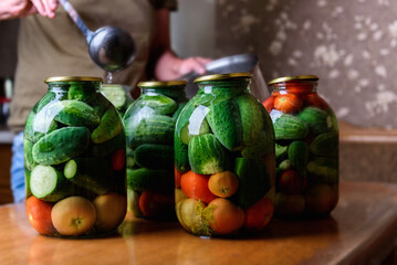 Preservation of season vegetables. A woman pouring the prepared marinade into jars with cucumbers and tomatoes in the kitchen.